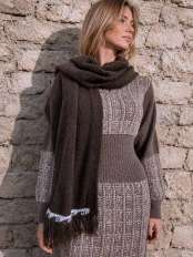 Femme blonde devant un mur en pierre, portant une robe en maille et une étole marron en laine d’alpaga à franges, modèle Kora.